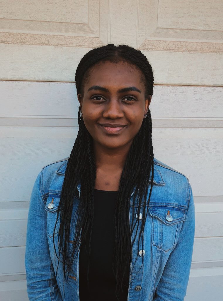 Toyo Ajibolade stands in front of a white wall, wearing a jean jacket and smiling at the camera.