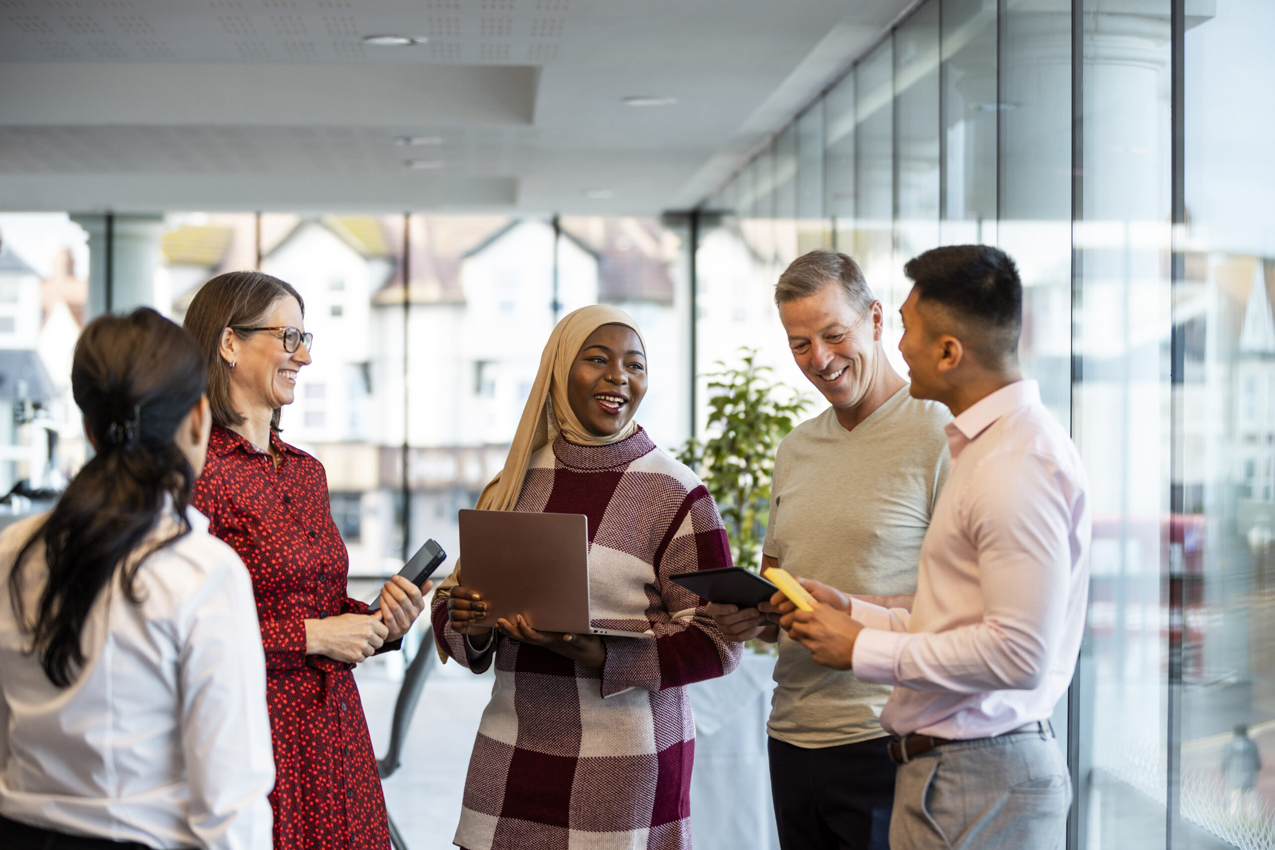 Medium shot of a mixed ethnic group of business professionals at a networking conference/meeting room in the North East of England. They are standing holding laptops/digital tablets smiling while discussing business together.