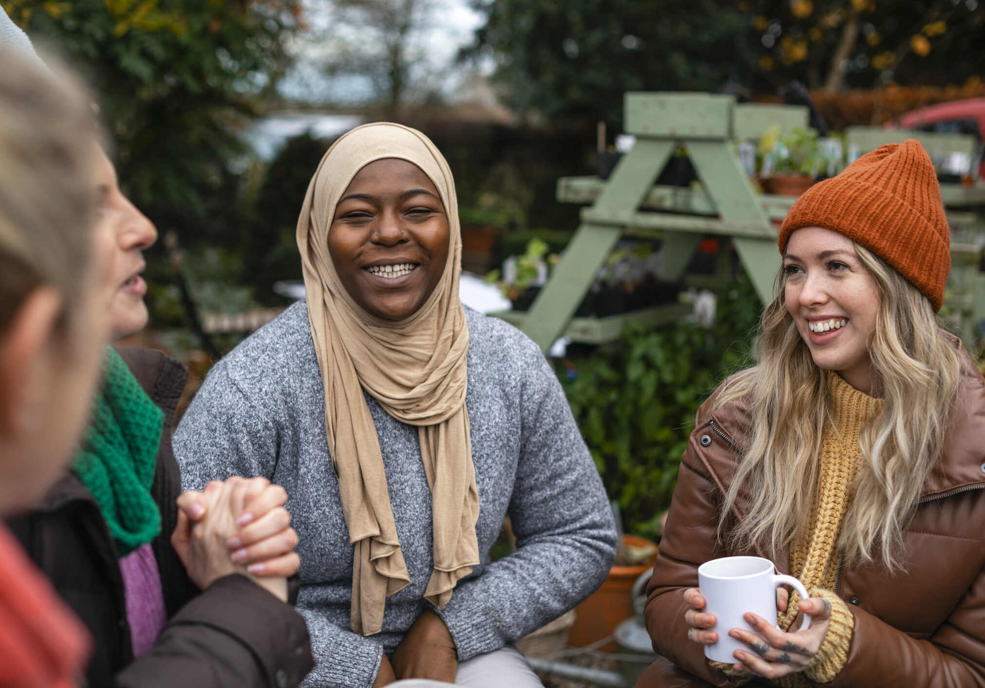 Volunteers sitting outdoors wearing warm casual clothing on a sunny cold winters day. They are resting and having a tea break from working on a community farm, looking after crops and performing other sustainable and environmentally friendly tasks. They are laughing and talking together, drinking hot drinks.